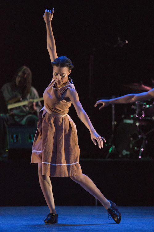 Karida Griffith in a organge dress stands on her right leg with her left toe dragging inwarm. Her arms make a diagonal; her right reaches high while her left reaches to the floor.
